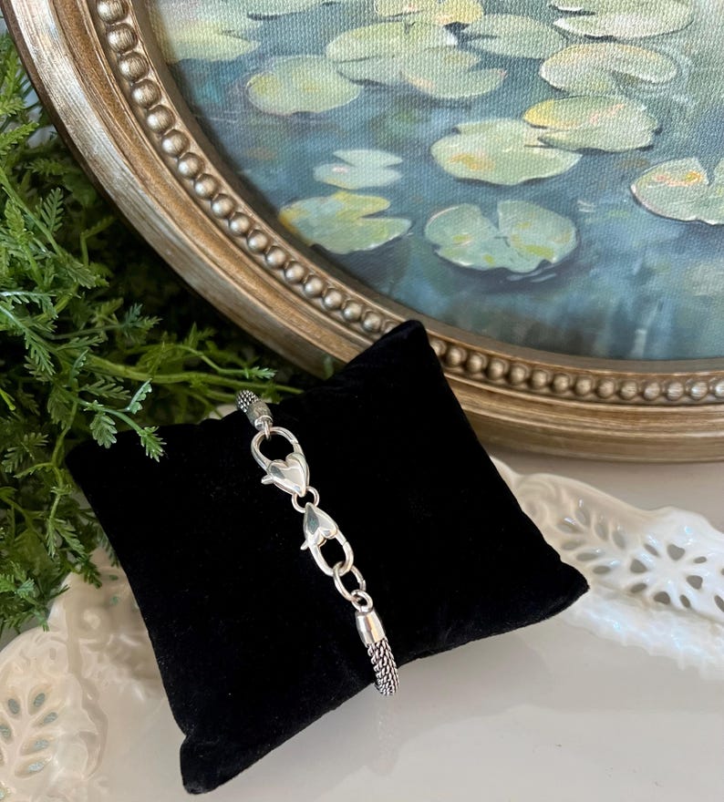 May include: A silver bracelet with a woven chain design and heart-shaped clasps is displayed on a black velvet pillow. The bracelet is shown against a backdrop of a framed painting of lily pads in a pond.