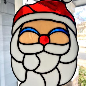 May include: A stained glass Santa Claus ornament with a red hat, white beard, and blue eyes. The ornament is hanging from a silver chain.