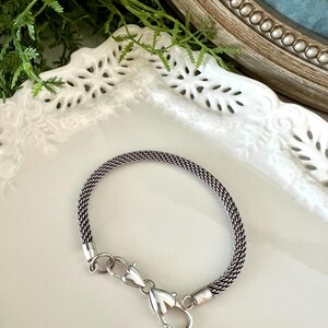 May include: A silver bracelet with a woven chain design. The bracelet has a lobster clasp closure. The jewellery is displayed on a white, decorative plate, with greenery in the background.