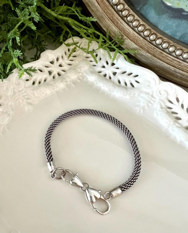 May include: A silver bracelet with a woven chain design. The bracelet has a lobster clasp closure. The jewellery is displayed on a white, decorative plate, with greenery in the background.