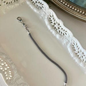 May include: A silver-coloured chain extender with a lobster clasp at one end and a small ring at the other. The chain has a woven texture. The extender is displayed on a white, decorative tray.