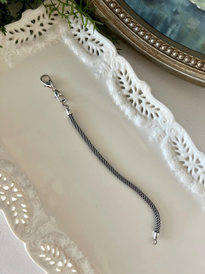 May include: A silver-coloured chain extender with a lobster clasp at one end and a small ring at the other. The chain has a woven texture. The extender is displayed on a white, decorative tray.