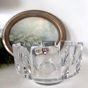 May include: A clear, faceted glass bowl with a scalloped rim. The bowl has a round base and a small, red and white label that reads "Orrefors Sweden". The bowl is displayed in front of an oval painting in a gold frame.
