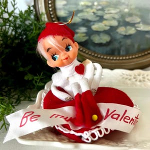 May include: A vintage Valentine's Day ornament featuring a doll with a red hat and dress, white hair, and blue eyes. The doll is holding a heart and sits on a heart-shaped base with a ribbon that reads "Be my Valentine."
