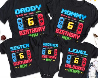 May include: Five black t-shirts and one black baby bodysuit featuring a pixelated video game console graphic with a yellow number 6 on the screen. The shirts display text including DADDY, MOMMY, SISTER, BROTHER, and LEVEL UNLOCKED HENRY.