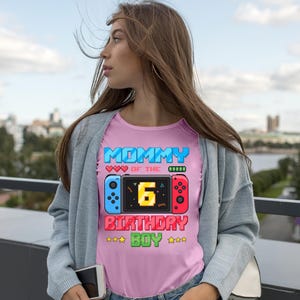 May include: Pink t-shirt with a pixelated design featuring the text "MOMMY OF THE 6 BIRTHDAY BOY." The design includes a game controller graphic with blue and red sides and a yellow number 6 in the center. The shirt is a crew neck.