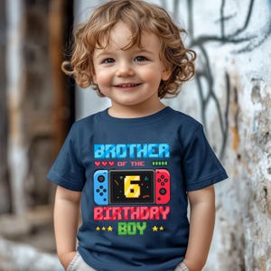 May include: Navy blue t-shirt with the text "BROTHER OF THE 6 BIRTHDAY BOY" in a pixelated font. The design includes a game controller graphic with blue and red accents. The shirt is for a child.