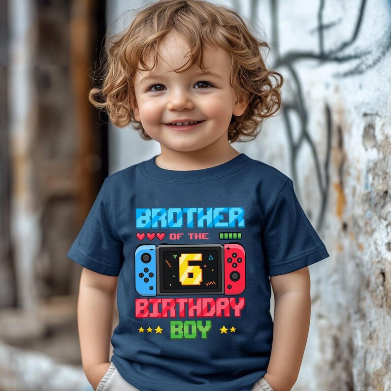 May include: Navy blue t-shirt with the text "BROTHER OF THE 6 BIRTHDAY BOY" in a pixelated font. The design includes a game controller graphic with blue and red accents. The shirt is for a child.