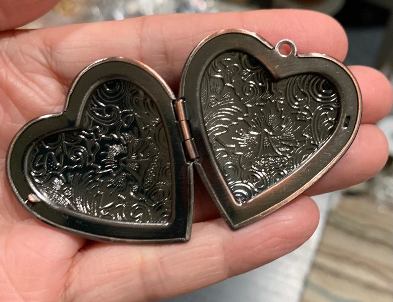 K&ouml;nnte beinhalten: Ein herzf&ouml;rmiges Medaillon mit einem detaillierten Blumenmuster auf beiden Seiten. Das Medaillon ist aus Metall mit einer kupfernen Oberfl&auml;che.