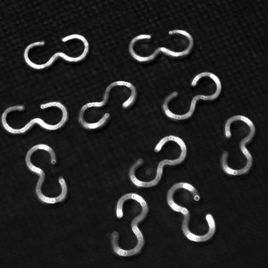 May include: A close-up of ten silver-toned jump rings, each shaped like a figure eight. The rings are scattered on a black surface.