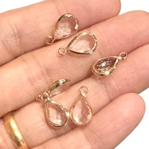 May include: Several teardrop-shaped charms with a clear, faceted center and a gold-colored metal frame. Each charm has a small loop at the top for attaching to jewelry. The charms are displayed against a neutral background.