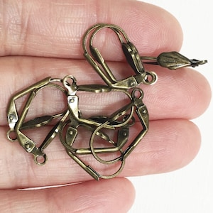 Puede incluir: Primer plano de un montón de ganchos para pendientes de color dorado antiguo. Los ganchos son de metal y tienen un cierre de resorte.