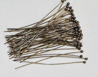 May include: A pile of brass-colored head pins with small, round heads. The pins are arranged in a loose cluster, with the pointed ends facing different directions. The background is a plain white surface. These pins are likely used for crafting or sewing projects.