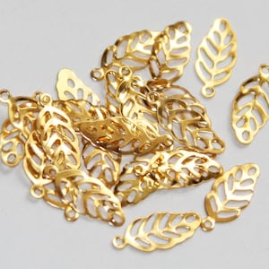 Op de afbeelding: Een groep gouden metalen bladcharms met een delicaat, opengewerkt ontwerp. Elke charme heeft een kleine lus aan de bovenkant om te bevestigen aan sieraden of andere knutselprojecten.