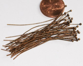 May include: A pile of copper-colored head pins with round heads. The pins are about 1 inch long and are arranged in a loose pile. A penny is in the background for size comparison.