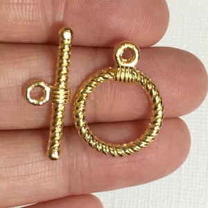 May include: Gold-tone metal toggle clasp with a twisted rope design. The clasp is made of two pieces: a bar with a loop on one end and a ring with a loop on top.