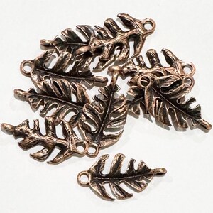 May include: A collection of bronze-colored leaf charms. Each charm features a detailed leaf design with visible veins and a small loop for attaching to jewelry or other crafts. The charms have a rustic, antique appearance.