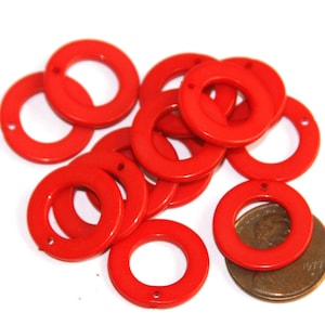 May include: A collection of bright red circular flat rings, each featuring a single small hole at the top. These circular components are arranged in a loose pile next to a copper-colored United States penny for scale, showing their relative size.
