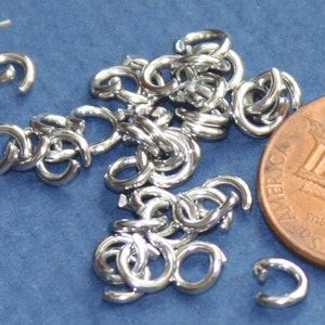 Puede incluir: Un montón de anillas de salto de plata, algunas están abiertas y otras están cerradas. Las anillas de salto están al lado de un centavo estadounidense.
