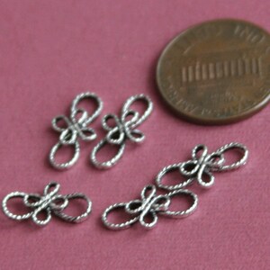 Op de afbeelding: Vijf zilveren metalen decoratieve knopen of sluitingen met een touwachtig ontwerp. De knopen zijn ongeveer zo groot als een Nederlandse cent.