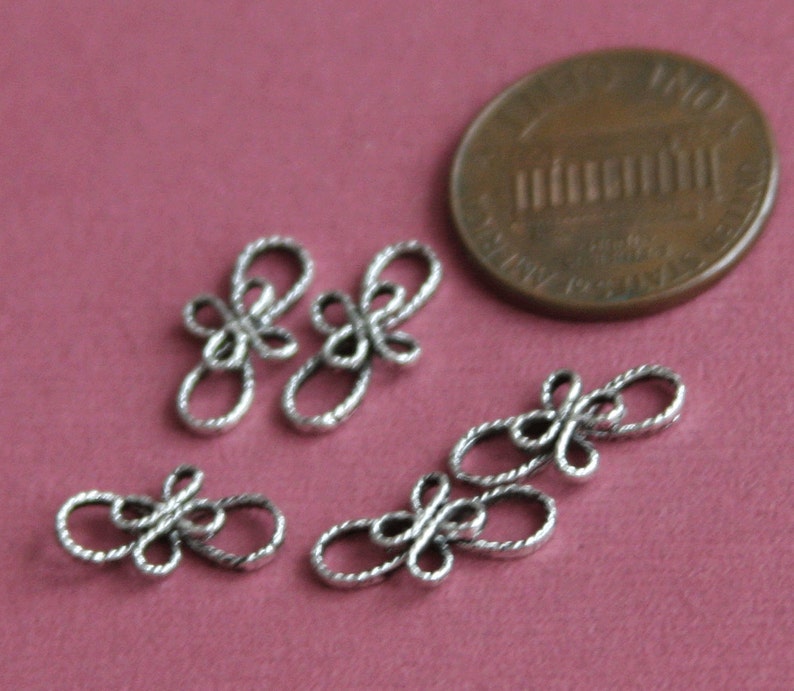 Op de afbeelding: Vijf zilveren metalen decoratieve knopen of sluitingen met een touwachtig ontwerp. De knopen zijn ongeveer zo groot als een Nederlandse cent.