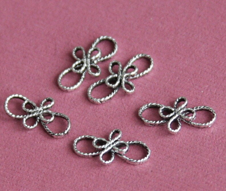 Op de afbeelding: Vijf zilverkleurige metalen decoratieve connectoren in de vorm van een gestileerde bloem met vier bloemblaadjes. De connectoren zijn ontworpen om te worden gebruikt bij het maken van sieraden of andere knutselprojecten.