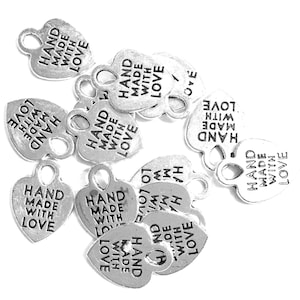 May include: A collection of silver-colored metal heart-shaped charms, each featuring a small loop at the top for attachment. The text "HAND MADE WITH LOVE" is embossed in black lettering on the front surface of every individual heart charm.