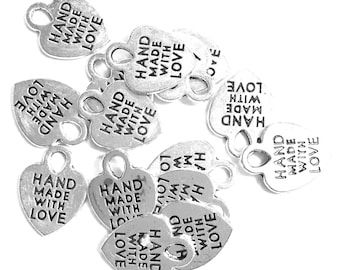 May include: A collection of silver-colored metal heart-shaped charms, each featuring a small loop at the top for attachment. The text "HAND MADE WITH LOVE" is embossed in black lettering on the front surface of every individual heart charm.