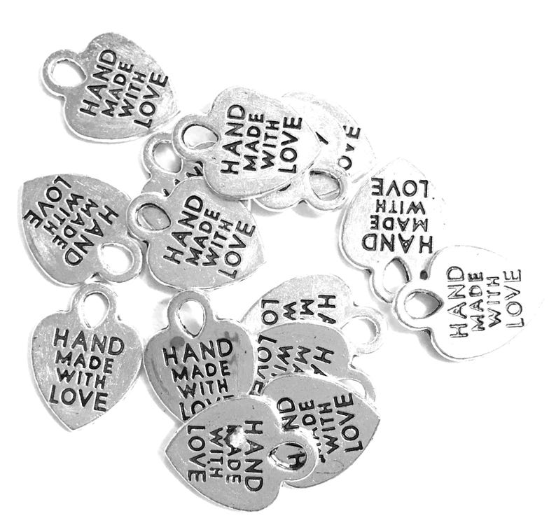 May include: A collection of silver-colored metal heart-shaped charms, each featuring a small loop at the top for attachment. The text "HAND MADE WITH LOVE" is embossed in black lettering on the front surface of every individual heart charm.