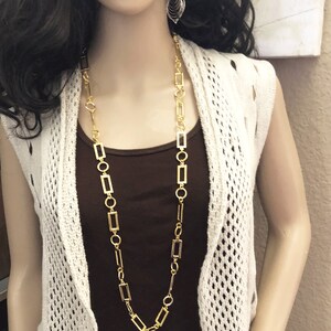 May include: A gold chain necklace with rectangular links. The necklace is long and hangs down to the chest. The necklace is worn by a mannequin with long dark hair and a white crocheted cardigan.