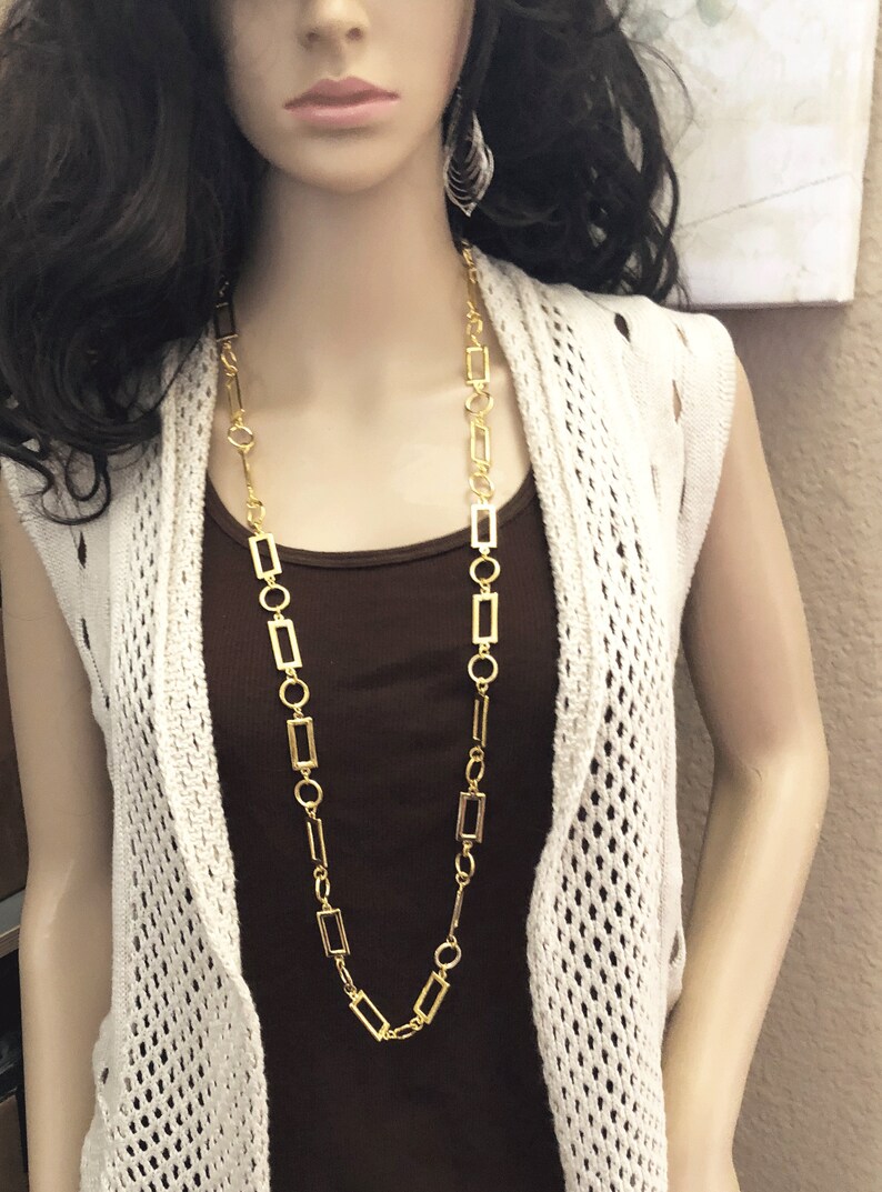 May include: A gold chain necklace with rectangular links. The necklace is long and hangs down to the chest. The necklace is worn by a mannequin with long dark hair and a white crocheted cardigan.