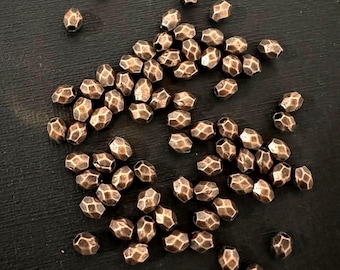 Könnte beinhalten: Eine Ansammlung kleiner, facettierter kupferfarbener Perlen mit metallischem Glanz. Jede Perle weist eine unregelmäßige, gehämmerte Textur und eine abgerundete, mehrflächige Form auf, die lose und ungeordnet im Bild verteilt sind.
