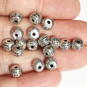 Puede incluir: Una colección de cuentas de metal plateadas con un diseño texturizado. Las cuentas son redondas y tienen un agujero en el centro para enhebrar.