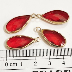 May include: Four teardrop-shaped red crystal pendants with gold-coloured metal edging. Each pendant has a small loop at the top for attaching to a chain or other jewellery component. The pendants are arranged on a white surface next to a ruler.