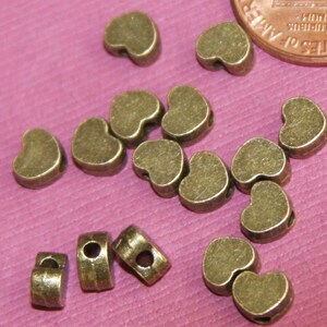 May include: A group of 14 antique gold heart-shaped beads with a hole through the centre. The beads are scattered on a pink surface.