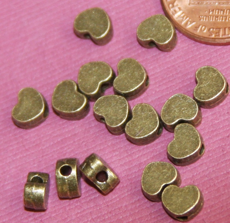 May include: A group of 14 antique gold heart-shaped beads with a hole through the centre. The beads are scattered on a pink surface.