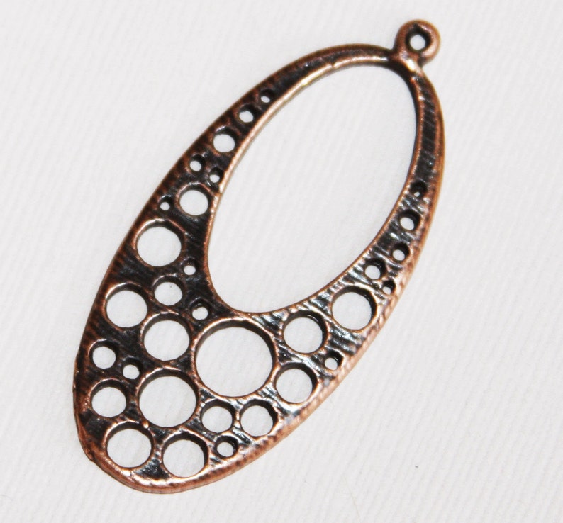 May include: A copper-colored oval pendant with a textured surface and a pattern of small circles cut out. The pendant has a loop at the top for hanging.