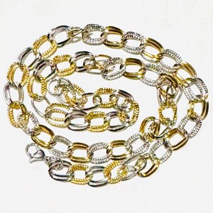 May include: A two-tone chain necklace with alternating gold and silver oval links. The necklace is displayed on a white surface, showcasing its intricate design and the contrast between the two metal colors. The clasp is visible at the end of the chain.