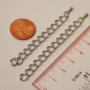 May include: Two silver chain extenders, each measuring approximately 3 inches in length, are laid out next to a ruler and a US penny for scale.
