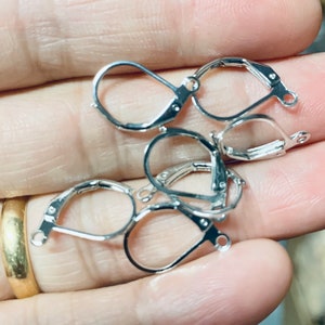 Puede incluir: Primer plano de seis ganchos para pendientes de plata, también conocidos como alambres para pendientes, para la elaboración de joyas. Los ganchos son de metal y tienen un acabado liso.