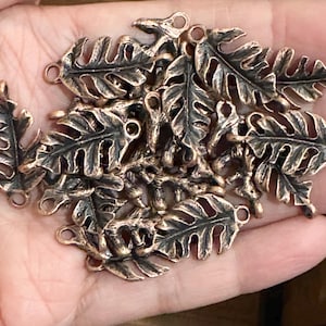 May include: A collection of antique copper-toned metal leaf charms, each featuring a detailed fern-like vein pattern and a small circular loop at the top and bottom for attachment. The charms have a dark, oxidized finish in the recessed areas.