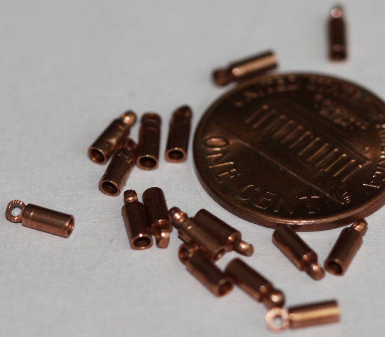 Puede incluir: Primer plano de un mont&oacute;n de tapones met&aacute;licos de color cobre para la fabricaci&oacute;n de joyas. Los tapones est&aacute;n esparcidos sobre una superficie blanca junto a una moneda de un centavo de d&oacute;lar estadounidense.