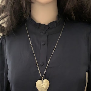 K&ouml;nnte beinhalten: Ein goldener herzf&ouml;rmiger Anh&auml;nger an einer zarten goldenen Kettenkette. Der Anh&auml;nger hat ein filigranes Design und wird auf einem schwarzen Hemd getragen.