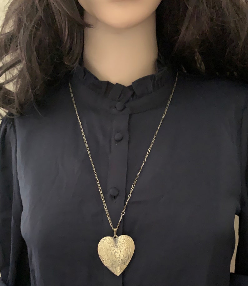 K&ouml;nnte beinhalten: Ein goldener herzf&ouml;rmiger Anh&auml;nger an einer zarten goldenen Kettenkette. Der Anh&auml;nger hat ein filigranes Design und wird auf einem schwarzen Hemd getragen.