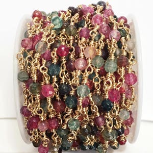 May include: A spool of gold chain with faceted gemstone beads in shades of pink, green, and brown. The beads are strung on a gold chain and are approximately 6mm in diameter.
