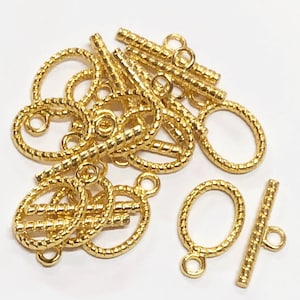 Puede incluir: Primer plano de un montón de cierres de palanca de metal dorado. Los cierres son de forma ovalada con un diseño de cuerda trenzada y un pequeño bucle en un extremo. También hay una barra recta con un diseño de cuerda trenzada y un bucle en cada extremo.