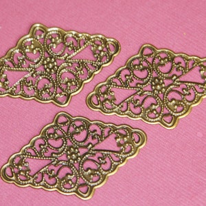 May include: Three gold-colored metal filigree embellishments in the shape of a diamond. The embellishments have a delicate, intricate design with a floral motif.