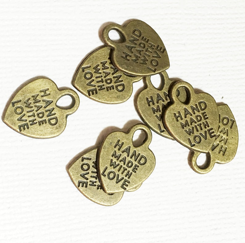 May include: A group of six gold heart-shaped metal tags with the text "HAND MADE WITH LOVE" on each tag. The tags have a hole at the top for attaching to a product.