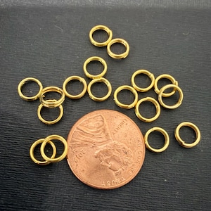 May include: A collection of small, gold-colored split rings, scattered on a dark surface. A copper penny is included for size comparison. The rings are likely used for jewelry making or crafts.