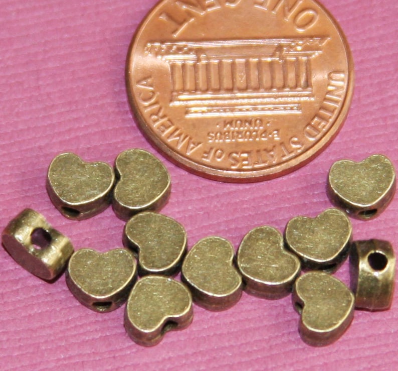 May include: A group of 10 antique gold heart-shaped metal beads with a hole in the centre. The beads are about the size of a UK penny.
